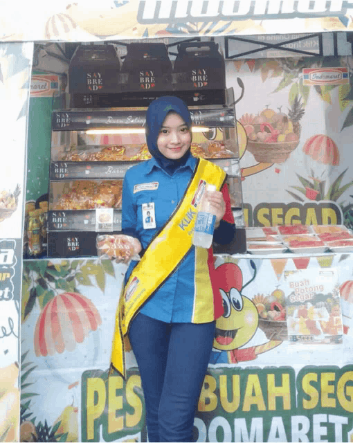 a woman in a blue shirt and yellow sash stands in front of a sign that says buah seg