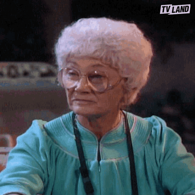 an elderly woman wearing glasses and a blue shirt is sitting at a table with a tv land logo in the corner .
