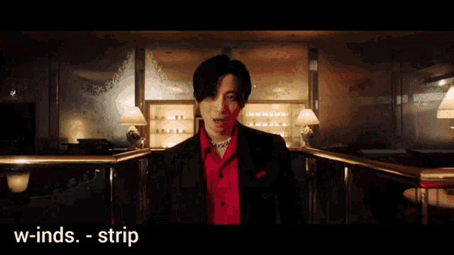 a man in a red shirt and black jacket is standing in a dark room with the words w-inds-strip below him