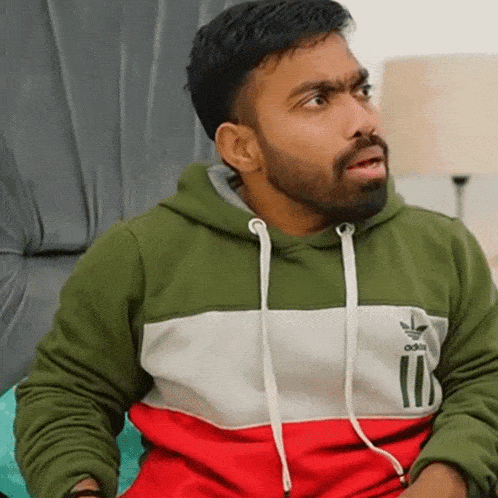 a man with a beard wearing a green and red adidas hoodie is sitting on a chair .