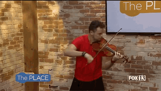 a man in a red shirt is playing a violin in front of a brick wall and a sign that says " the place "
