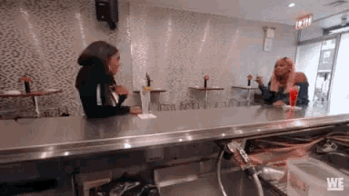 two women are sitting at a counter in a restaurant with a sign that says we on it .