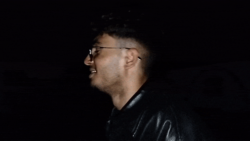 a young man wearing glasses and a black leather jacket is smiling in the dark .