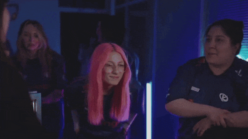 a woman with pink hair is standing in a dark room with other women .