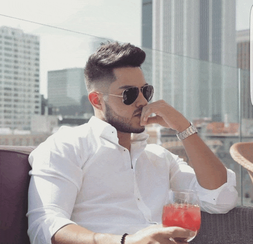 a man in a white shirt and sunglasses holds a glass of red liquid