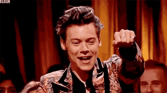 harry styles is wearing a floral suit and tie while giving a thumbs up in front of a crowd .