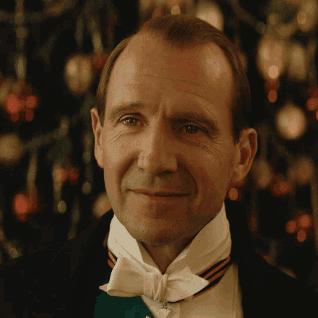 a man in a tuxedo with a bow tie smiles in front of a christmas tree