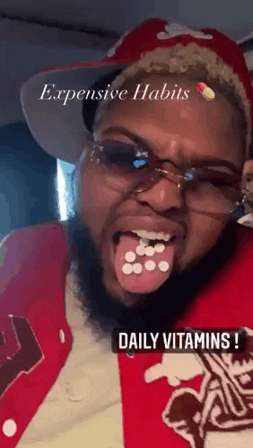 a man with a beard wearing glasses and a red jacket is holding pills in his mouth .