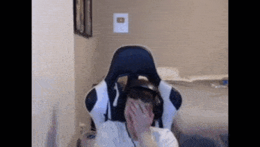 a man wearing headphones is sitting in a gaming chair and covering his face with his hand .