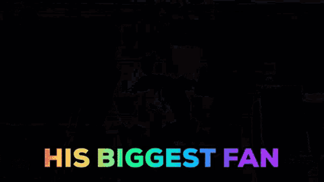 a man stands on a stage in front of a crowd with the words his biggest fan above him