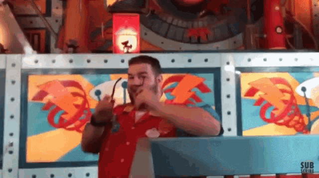 a man in a red shirt is giving a thumbs up in front of a colorful wall with the word sub written on it