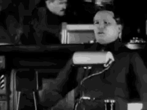 a black and white photo of a man sitting at a table talking on a phone .