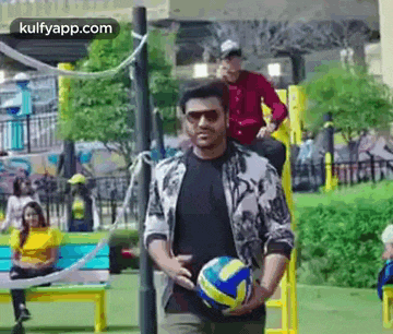 a man is holding a volleyball in his hands in a park while a woman sits on a bench .
