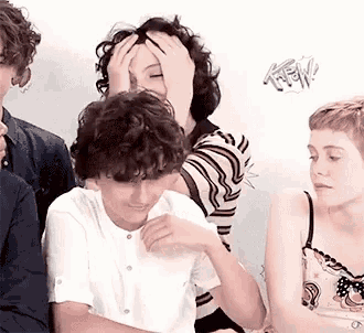 a group of young people are sitting in front of a wall and one of them is covering her face with her hand .