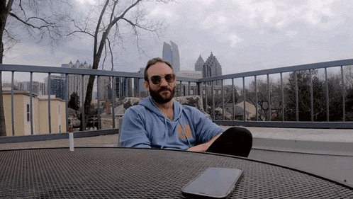 a man wearing sunglasses sits at a table with a cell phone and a cigarette