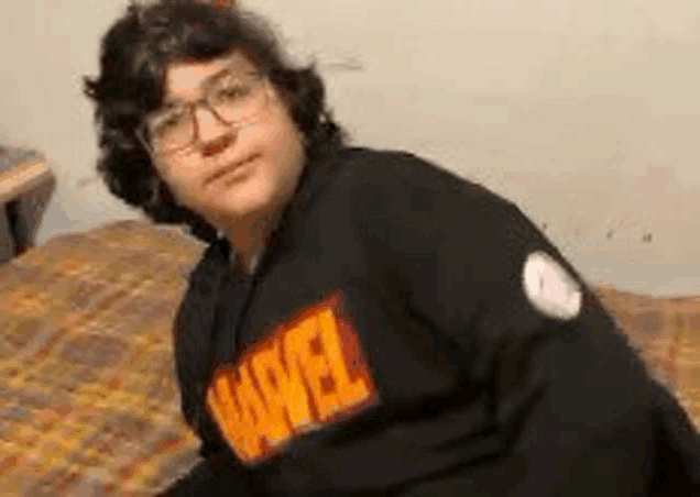 a young man wearing glasses and a black marvel sweatshirt is sitting on a bed .