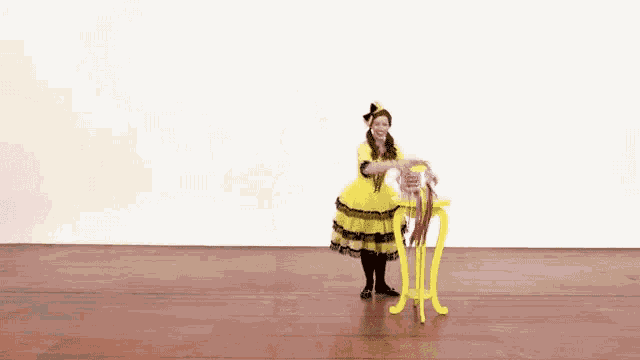 a woman in a yellow dress is dancing in front of a yellow table with a pink box on it