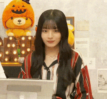 a young woman is sitting in front of a computer in a room with a stuffed animal in a pumpkin hat .
