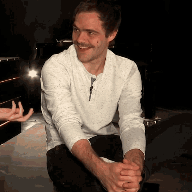 a man in a white shirt is smiling and sitting with his hands folded