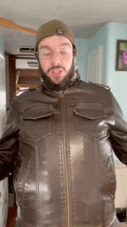 a man with a beard and glasses is wearing a brown leather jacket and a green beanie .