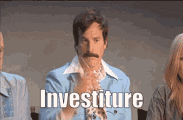 a man with a mustache is sitting in front of a group of people and says " investiture "