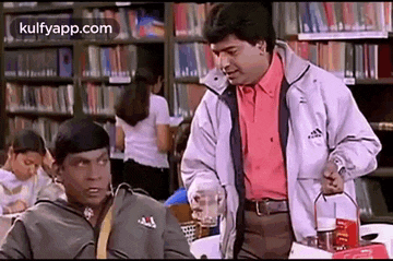 two men are standing in a library talking to each other . one of the men is holding a blender .