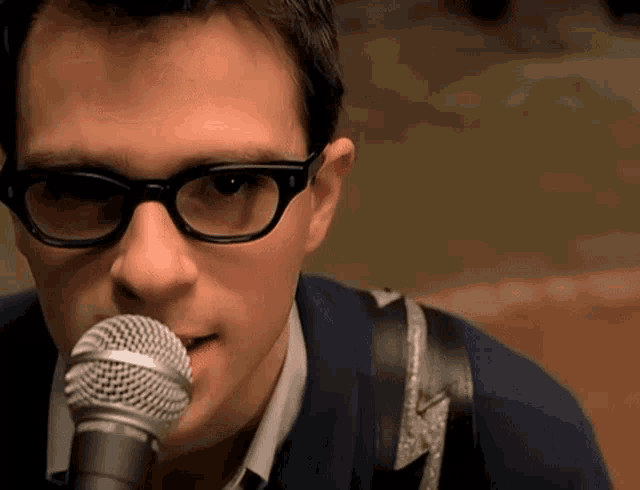a man wearing glasses sings into a shure microphone