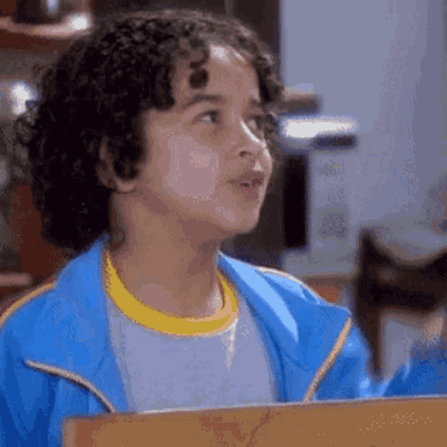 a young boy with curly hair wearing a blue jacket and a yellow t-shirt is sitting at a table .