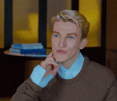 a young man with blonde hair and blue eyes is wearing a brown sweater and a blue striped shirt .