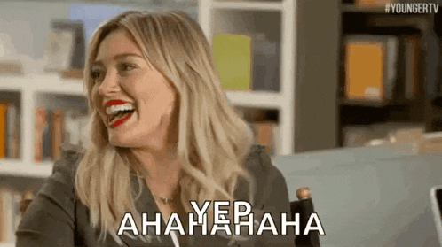 a woman is laughing while sitting in a chair in front of a bookshelf in a room .