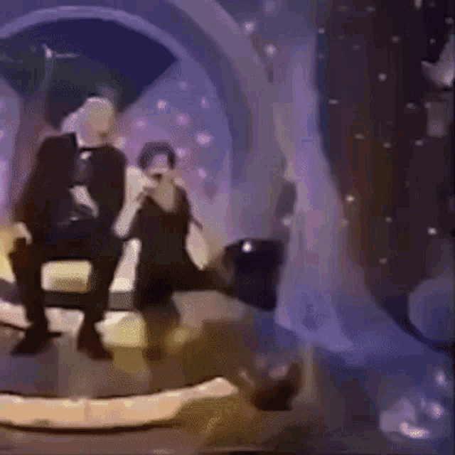 a man in a tuxedo sits next to a woman holding a microphone on a stage