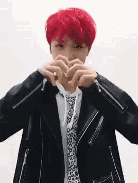a man with red hair is wearing a leather jacket and tie and making a heart shape with his hands .