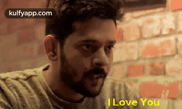 a man with a beard is sitting at a table in front of a brick wall and says `` i love you '' .