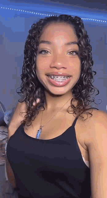 a young woman with curly hair and braces on her teeth is wearing a black tank top and a necklace .