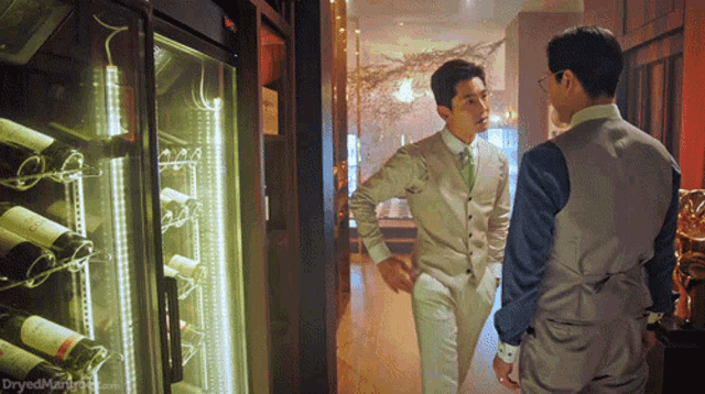 two men in suits are standing in front of a wine cooler with bottles of wine on it