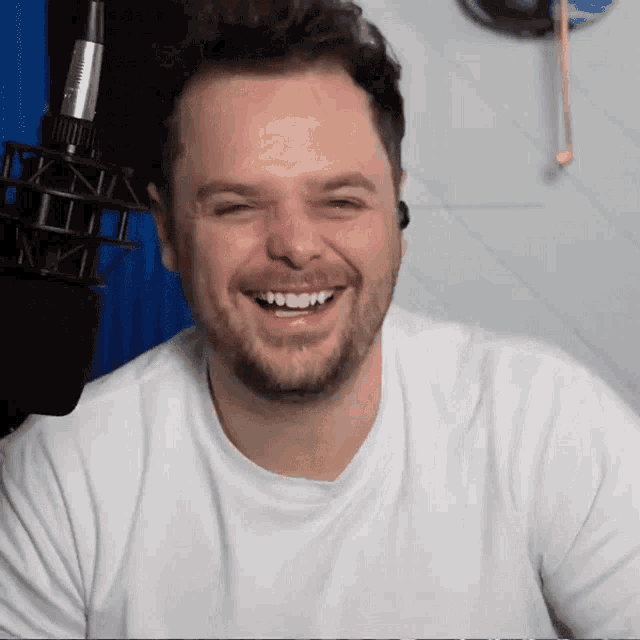 a man with a beard wearing a white shirt is smiling in front of a microphone