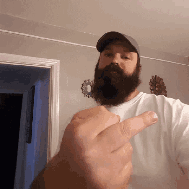 a man with a beard wearing a baseball cap and a white shirt giving the middle finger