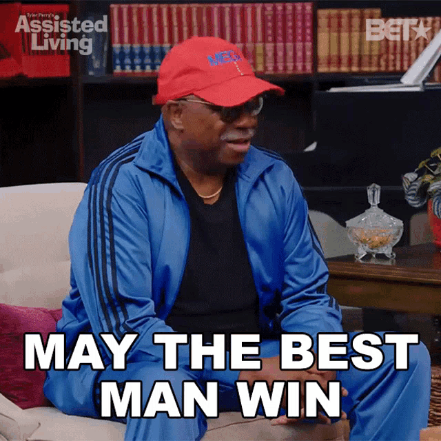 a man in a blue jacket and red hat is sitting on a couch and says may the best man win