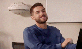 a man in a blue sweater is sitting at a desk with a laptop and smiling .