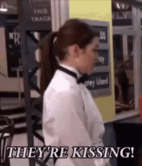 a woman in a white shirt and bow tie is standing in front of a sign that says `` they 're kissing '' .