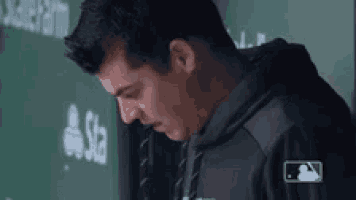 a man wearing a black hoodie is standing in front of a green wall with a mlb logo on it .