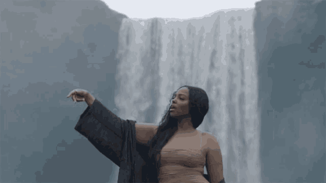 a woman stands in front of a waterfall wearing a black robe