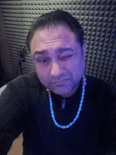 a man wearing a black sweater and a blue necklace has his eye closed
