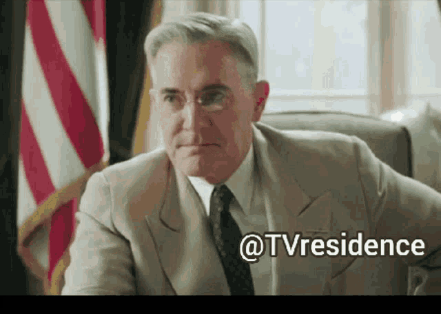 a man in a suit and tie is sitting in front of an american flag and the words @tvresidence are below him