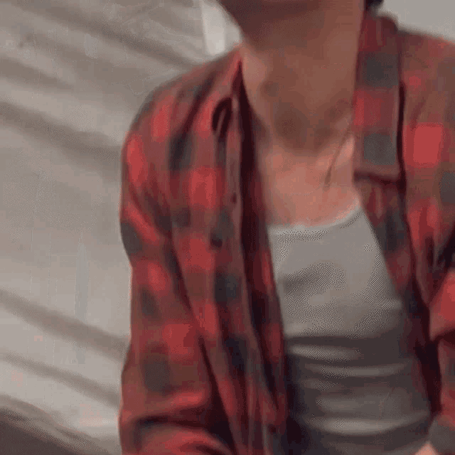 a man wearing a plaid shirt and a grey tank top is sitting on a bed .