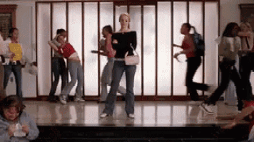a group of girls are dancing in a hallway with a woman standing in the middle .