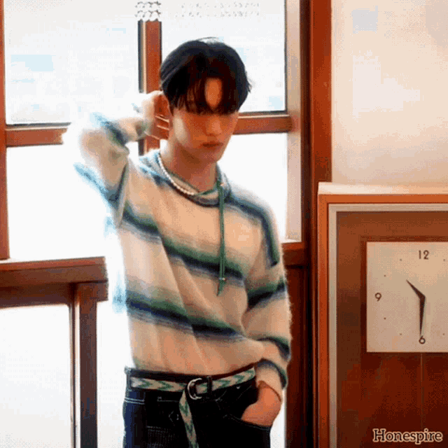 a man in a striped sweater stands in front of a clock that shows the time as almost 5:00