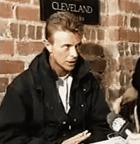 a man in a black jacket is sitting at a table in front of a brick wall .