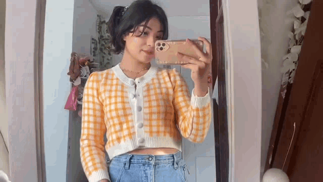 a woman is taking a selfie in front of a mirror while wearing a plaid sweater and jeans .