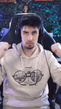 a man wearing headphones and a picture hoodie is sitting in a chair with his fist in the air .